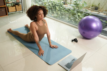 Hispanic woman working out on yoga mat at home, following tutorial on laptop