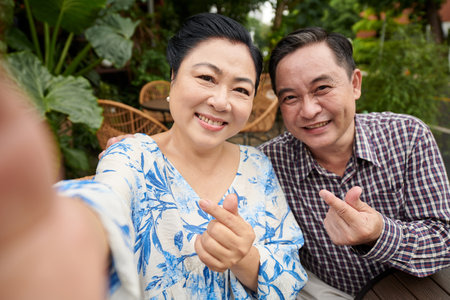 Vietnamese senior couple making finger hearts when taking selfie outdoorsの写真素材