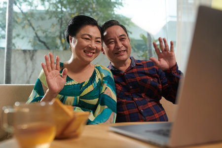 Happy senior Vietnamese couple waving with hands when video calling family membersの写真素材