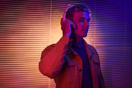 Portrait of young man standing in neon light and listening to his favorite music track in headphonesの写真素材