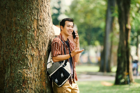 Smiling young man leaning on tree when talking on phone with friendの写真素材