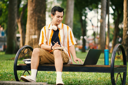 Smiling student sitting on bench in park and working on project for university class