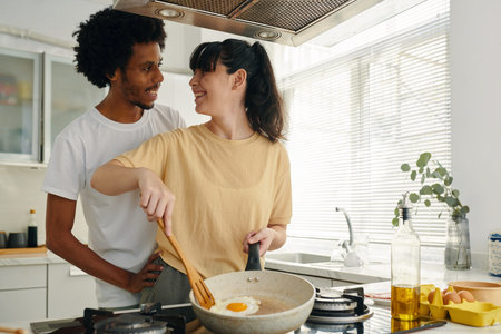 Pretty young woman with frying pan cooking scrambled eggs for breakfast while standing by electric...