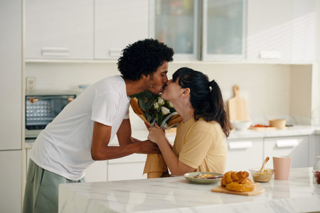 Young man passing flowers to his wife and kissing her on lips while congratulating his sweetheart on...