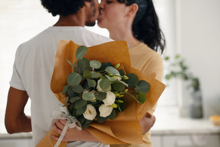 Bunch of beautiful white flowers wrapped in paper held by young amorous woman embracing her husband during passionate kissの写真素材