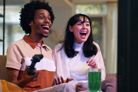 Young excited intercultural husband and wife sitting on couch in front of TV set and playing video...