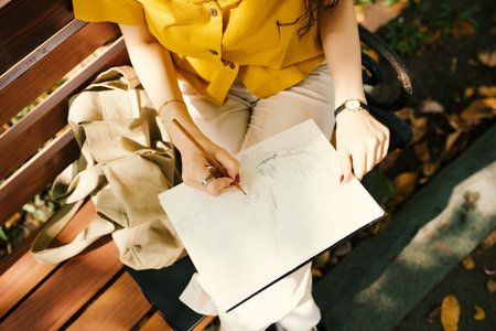 Above view of young woman in casual attire sitting on bench in park on weekend and drawing sketch of...