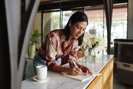 Female entrepreneur having morning coffee in coffeeshop and writing down ideas in plannerの写真素材