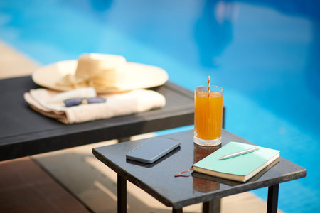 Glass of juice, planner and smartphone on table next to swimming poolの写真素材