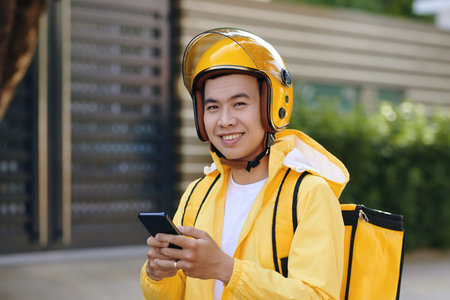 Portrait of smiling Vietnamese delivery man in yellow uniform checking orders from customers via...