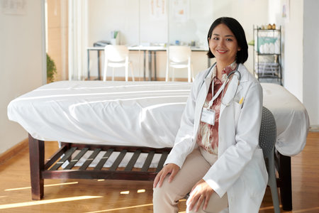 Smiling general practitioner sitting in front of hospital bed in wardの写真素材