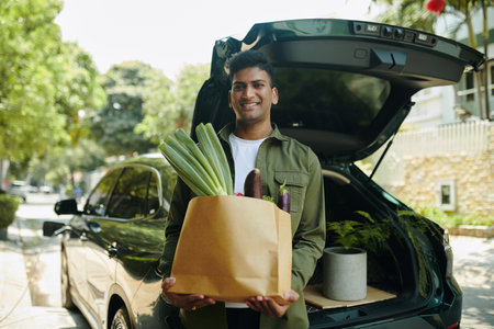 Portrait of smiling Indian man standing at opened car trunk with bag of groceriesの写真素材