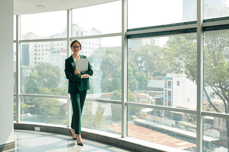Portrait of female entrepreneur with laptop standing at panoramic office windowの写真素材