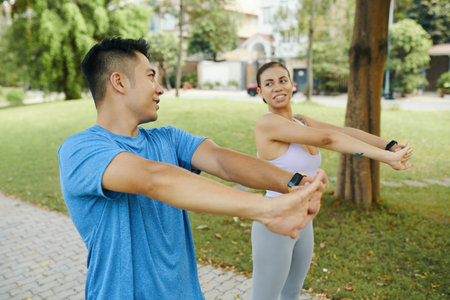 Man and woman in athletic wear warming up together by stretching their arms during an outdoor...