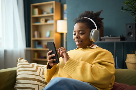 Woman wearing headphones, listening to music and holding smartphone while sitting on a cozy sofa in...