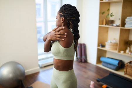 Young woman performing shoulder stretch in a minimalist home gym with wooden shelving in backgroundの写真素材