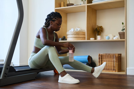African American woman wearing athletic wear, sitting at exercise area, holding water bottle and...