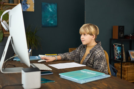 Portrait of young student working on computer at home. The student is sitting at wooden desk with various books and stationery, looking focusedの写真素材