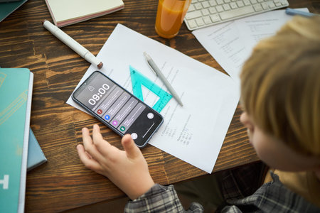 Child engaging in study session with smartphone, pen, paper, and books on wooden table, appearing...