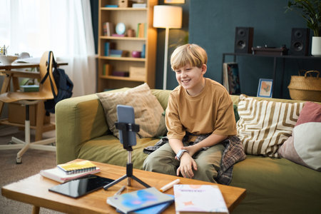 Blond boy sitting on couch in cozy living room with books and a phone on table, smiling and wearing casual attire, looking towards smartphone cameraの写真素材