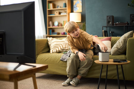 Young boy adjusting smart speaker while sitting on a green sofa in a cozy living room with bookshelves, cushions and small lamp in backgroundの写真素材