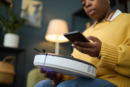 Woman sitting in living room, using smartphone to operate smart vacuum cleaner, lamp glowing warmly...