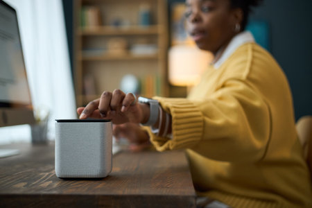 Black woman adjusting smart speaker on desk in cozy home office, with shelves and computer in background blending modern technology with comfortの写真素材