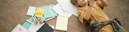 Young boy relaxing on carpet among scattered school supplies while using a device. Casual moment...