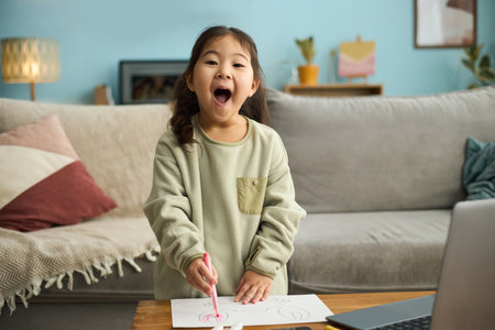 Young girl smiling brightly drawing with pink crayon on piece of paper in cozy living room featuring pillows, wooden furniture, and laptop on tableの写真素材