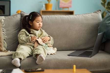 Little girl sitting on a couch holding stuffed toys while watching something on laptop with calm expressionの写真素材