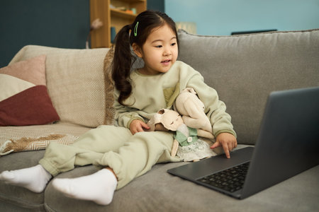 Asian girl sitting on couch, watching laptop screen while holding stuffed animals, cozy living room setting with child engrossed in screenの写真素材