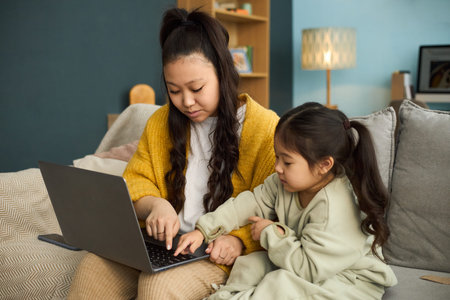 Multiracial mother and daughter engaging in learning activities on laptop in cozy living room,...