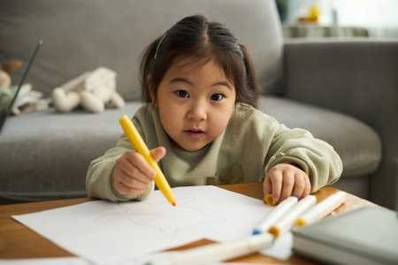 Portrait of young Asian girl engaging in drawing activity, holding a yellow crayon with focused expressionの写真素材
