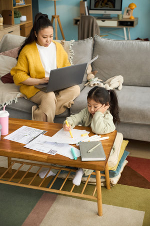 Woman on gray couch using laptop while young girl working on school assignments at wooden coffee table, surrounded by papers and booksの写真素材