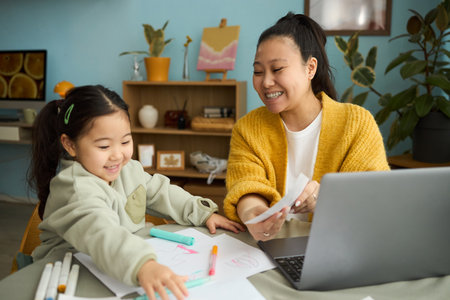 Smiling Asian mother sharing a joyful moment with daughter while doing a creative art project together at home on a cozy afternoonの写真素材