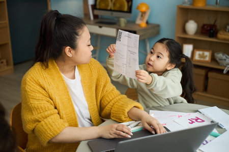 Mother assisting young daughter with schoolwork at table, surrounded by learning materials and books, they both focusing on documentの写真素材