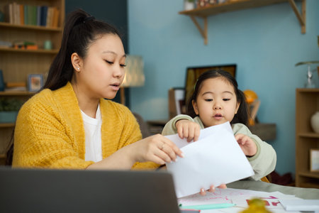 Mother assisting daughter with homework while sitting at desk at home, both focused on task at handの写真素材