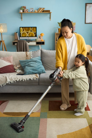 Mother in yellow cardigan and daughter in green sweatsuit vacuuming colorful living room with modern decor, shelves, and artworkの写真素材