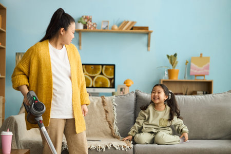 Mother in yellow cardigan using vacuum cleaner while looking at daughter sitting on couch in cozy living room with shelves and wall decorationsの写真素材