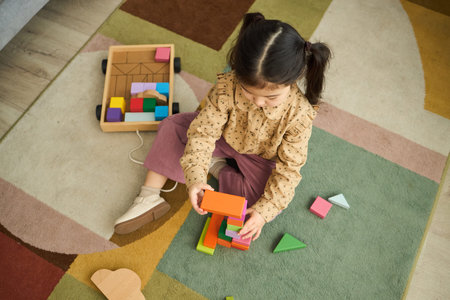 Child sitting on colorful rug building structures with wooden blocks while surrounded by other toys and patterned floorの写真素材