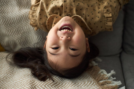 Portrait of young girl with long dark hair smiling, while lying upside down on soft couch with patterned cushions and knit blanket surrounded by cozy home decorの写真素材