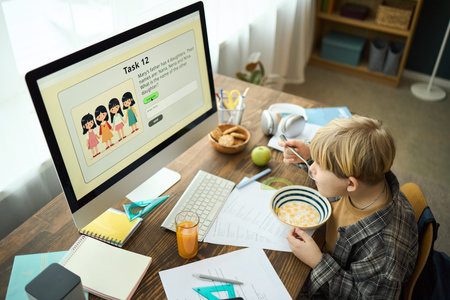 Young boy participating in online learning session while having breakfast at home. Workspace with...