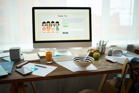 Computer with digital elementary school homework on screen, on desk with gadgets, papers, and breakfast items, natural light streaming through windowの写真素材