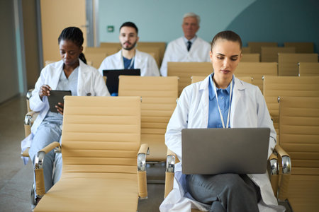 Group of medical professionals engaging with their laptops in hospital waiting room. Scene includes...