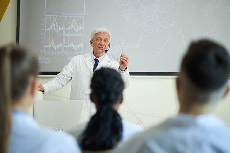 Senior scientist presenting research findings to attentive students in modern classroom environment with graphs displayed on the screen while making important pointsの写真素材