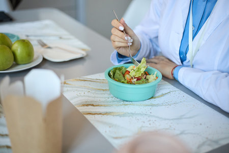 Medical professional taking a lunch break, eating healthy salad at office table, with fruits and utensils visible in backgroundの写真素材