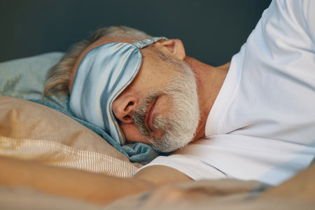 Elderly man wearing a sleep mask while resting peacefully on bed with pillow and blanket, capturing a serene moment of relaxationの写真素材