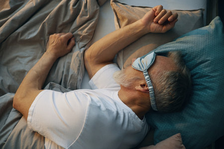 Elderly Caucasian man sleeping in bed with a sleep mask covering his eyes, resting peacefully while lying on his side with pillows and bedding surrounding himの写真素材