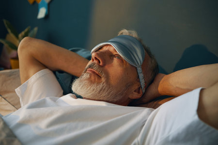 Senior man reclining on bed, wearing sleep mask and looking serene. Background shows minimalistic...