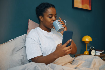 Young African American woman sipping tea while interacting with phone in cozy bedroom displaying...